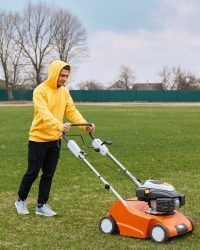 young attractive man trimming grass with cutter, young adult male wearing yellow hoodie and black trousers mowning lawn, pushing special equipment for taking care of field.
